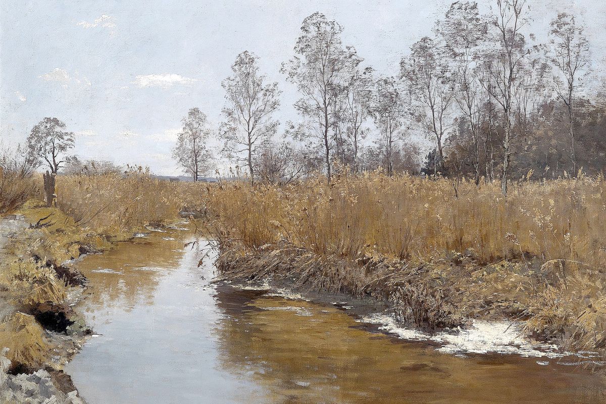 A slow stream bordered by dry golden reeds and leafless trees under a pale gray sky with patches of melting snow on the muddy